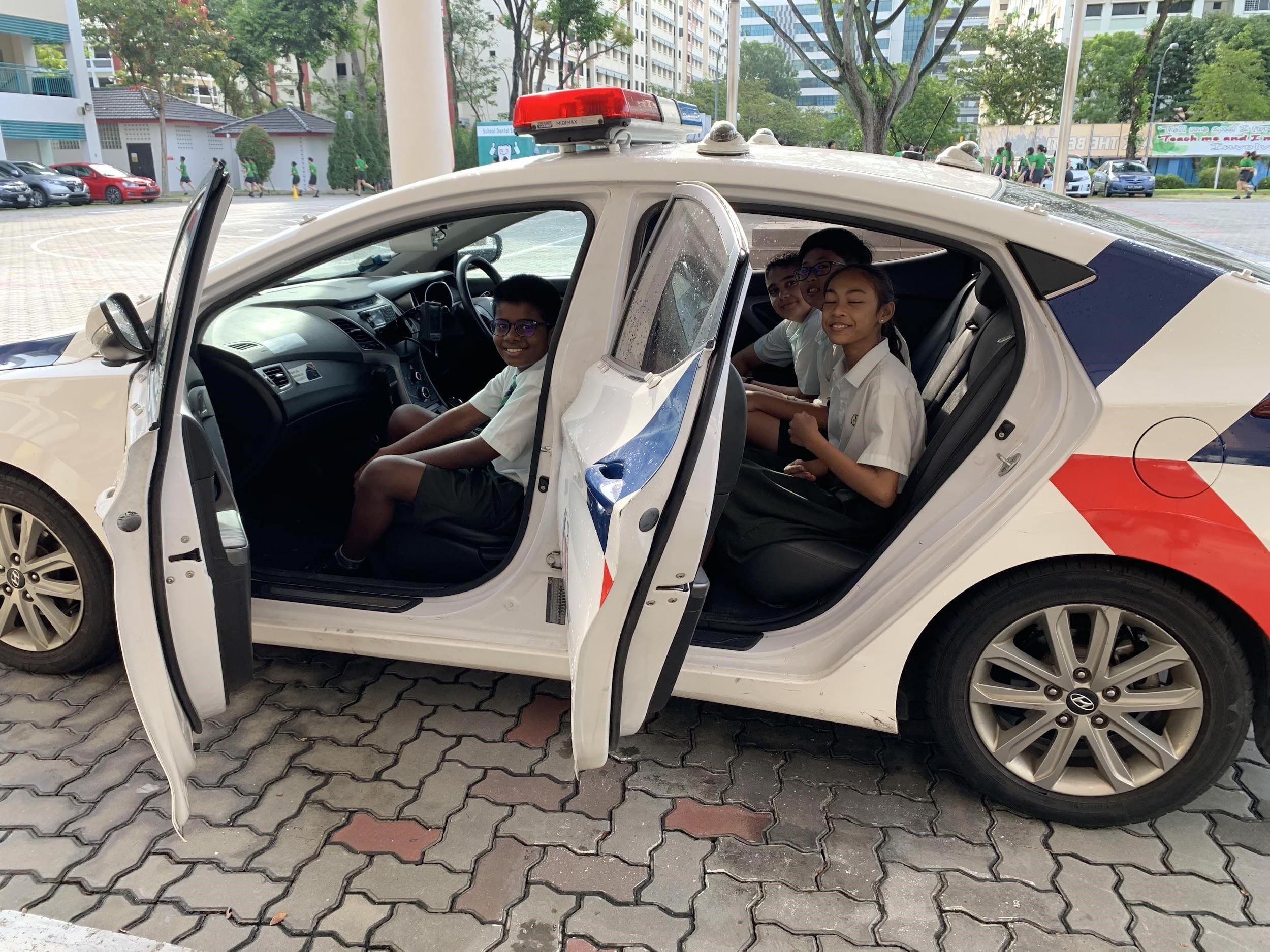 Three smiling school children sit inside a white police car with red and blue markings and a roof siren.